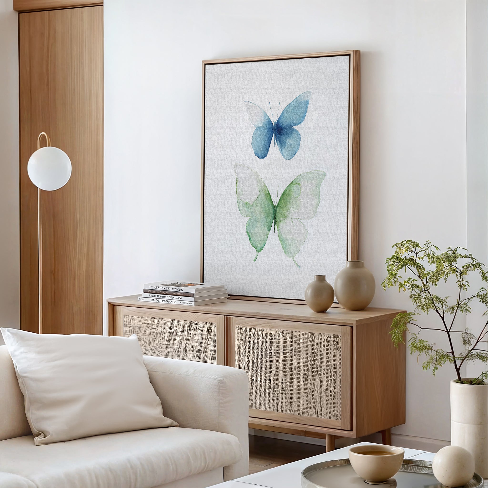 A modern living room with a light wood sideboard, neutral decor, and Soft Flight Canvas Art featuring blue and green watercolor butterflies. A white sofa and sleek table are in the foreground, with a potted plant and books on the sideboard.