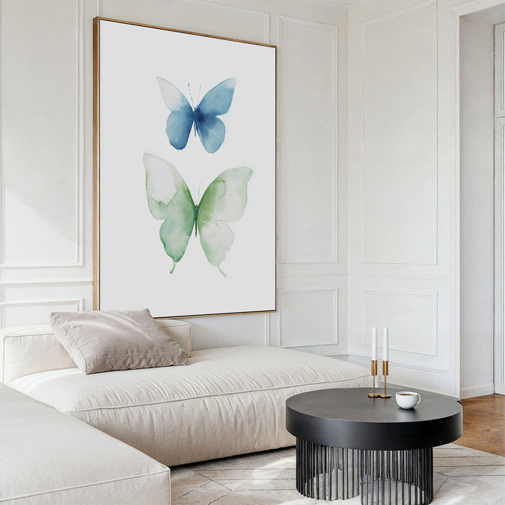 A modern living room with a white sectional, beige pillow, round black coffee table, and the Soft Flight Canvas Art—large minimalist wall art featuring blue and green watercolor butterflies for a Nordic Park-inspired look.