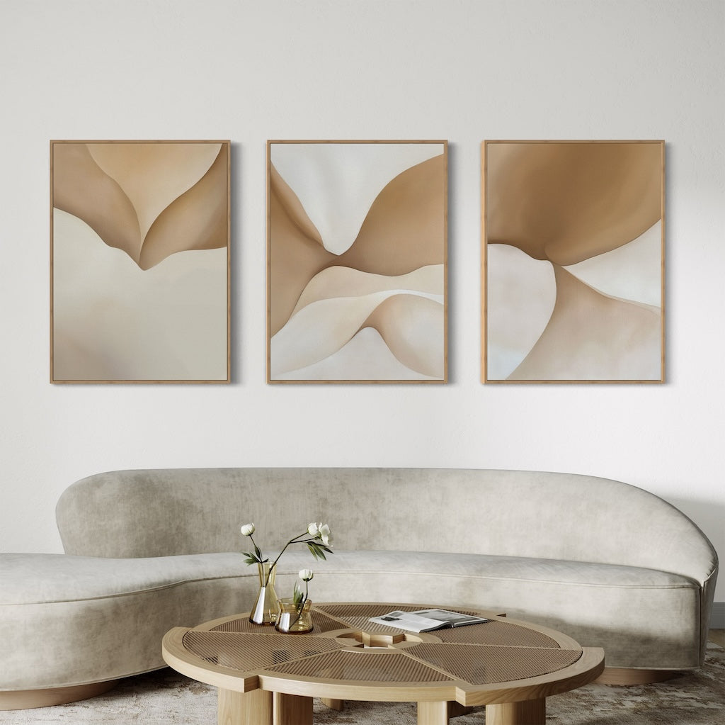 A modern living room features a curved beige sofa, a round wooden coffee table with a vase and magazines, and the Soft Sands Set of 3 abstract beige and brown paintings on the white wall for contemporary home décor.