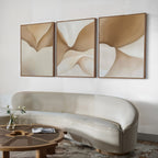 A modern living room with a curved beige sofa, a round wooden coffee table holding a vase and magazine, and the Soft Sands Set of 3 abstract beige and brown paintings displayed above the sofa.