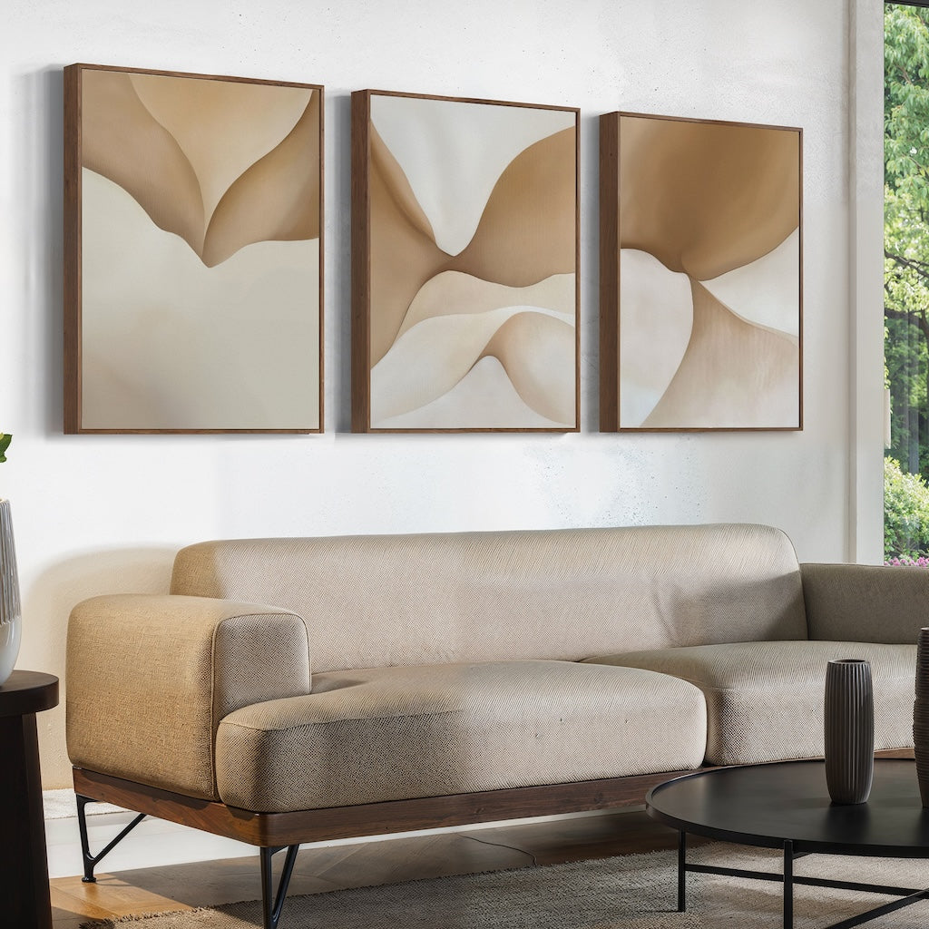 A modern living room features a beige sectional sofa, a round black coffee table, and the Soft Sands Set of 3 abstract wall art above the sofa. A window reveals greenery outside, adding to the contemporary décor.
