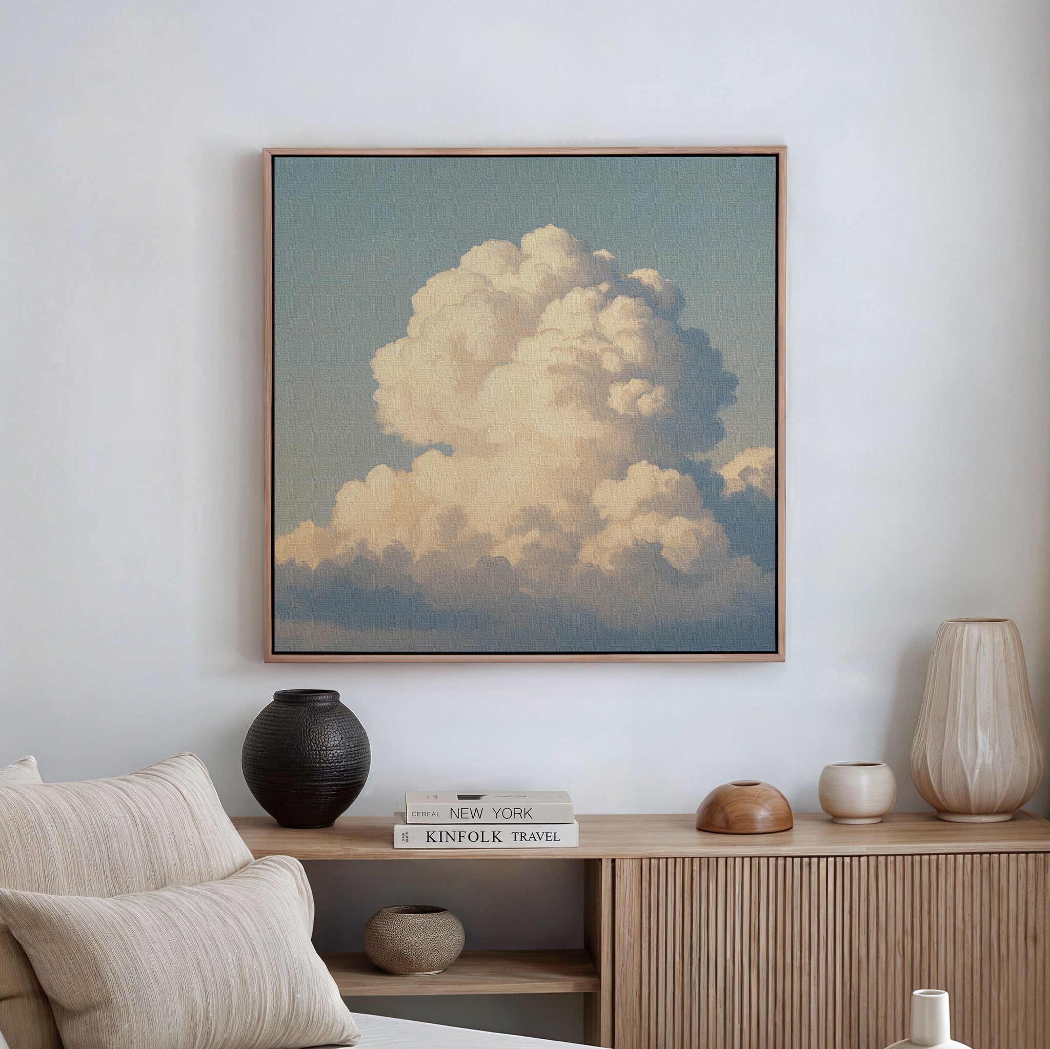 The Soft Skies Rising Canvas Art, featuring fluffy white clouds against a blue sky, is framed and displayed above a wooden sideboard with vases and books, near a low beige sofa on a white wall.
