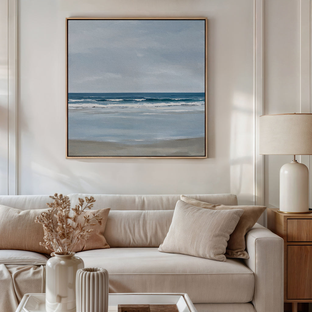 A modern living room with a white sofa, beige cushions, side table and lamp, dried flowers in a vase, and coastal decor featuring the Soft Tide Canvas Art of a calm ocean scene on the wall.