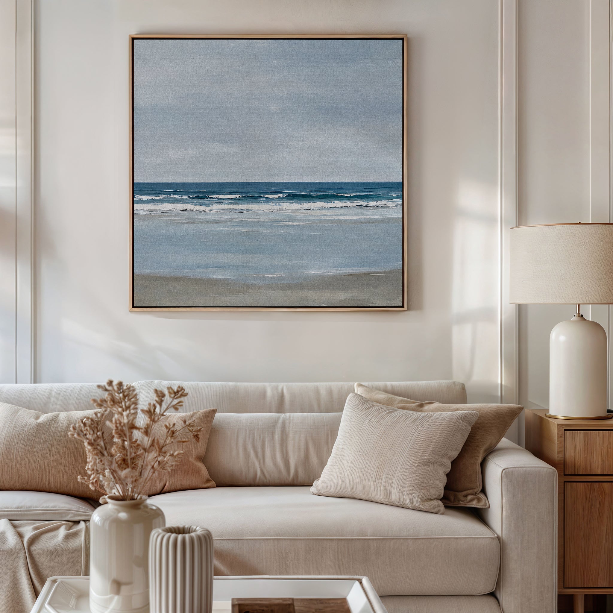 A modern living room with a white sofa, beige cushions, side table and lamp, dried flowers in a vase, and coastal decor featuring the Soft Tide Canvas Art of a calm ocean scene on the wall.