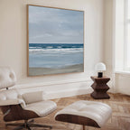 A modern, minimalist room with the Soft Tide Canvas Art as a large ocean landscape, a white leather lounge chair and ottoman, wooden side table, and small white lamp by a bright window—an ideal coastal decor accent.