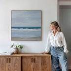 A woman in a white shirt and jeans stands by a wooden cabinet, admiring the Soft Tide Canvas Art—a framed seascape painting. A small plant and a closed book sit on the cabinet, enhancing the serene coastal decor.