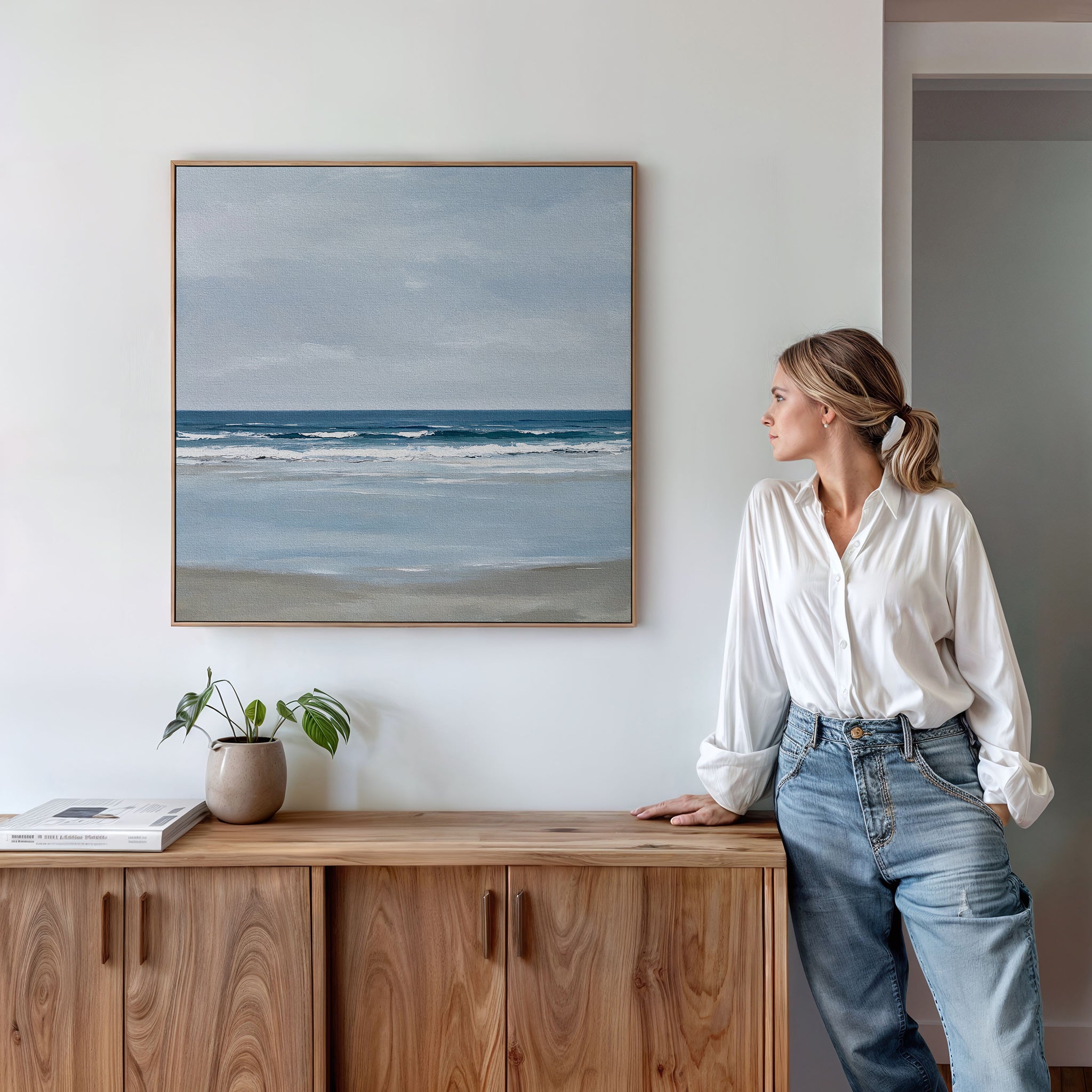 A woman in a white shirt and jeans stands by a wooden cabinet, admiring the Soft Tide Canvas Art—a framed seascape painting. A small plant and a closed book sit on the cabinet, enhancing the serene coastal decor.