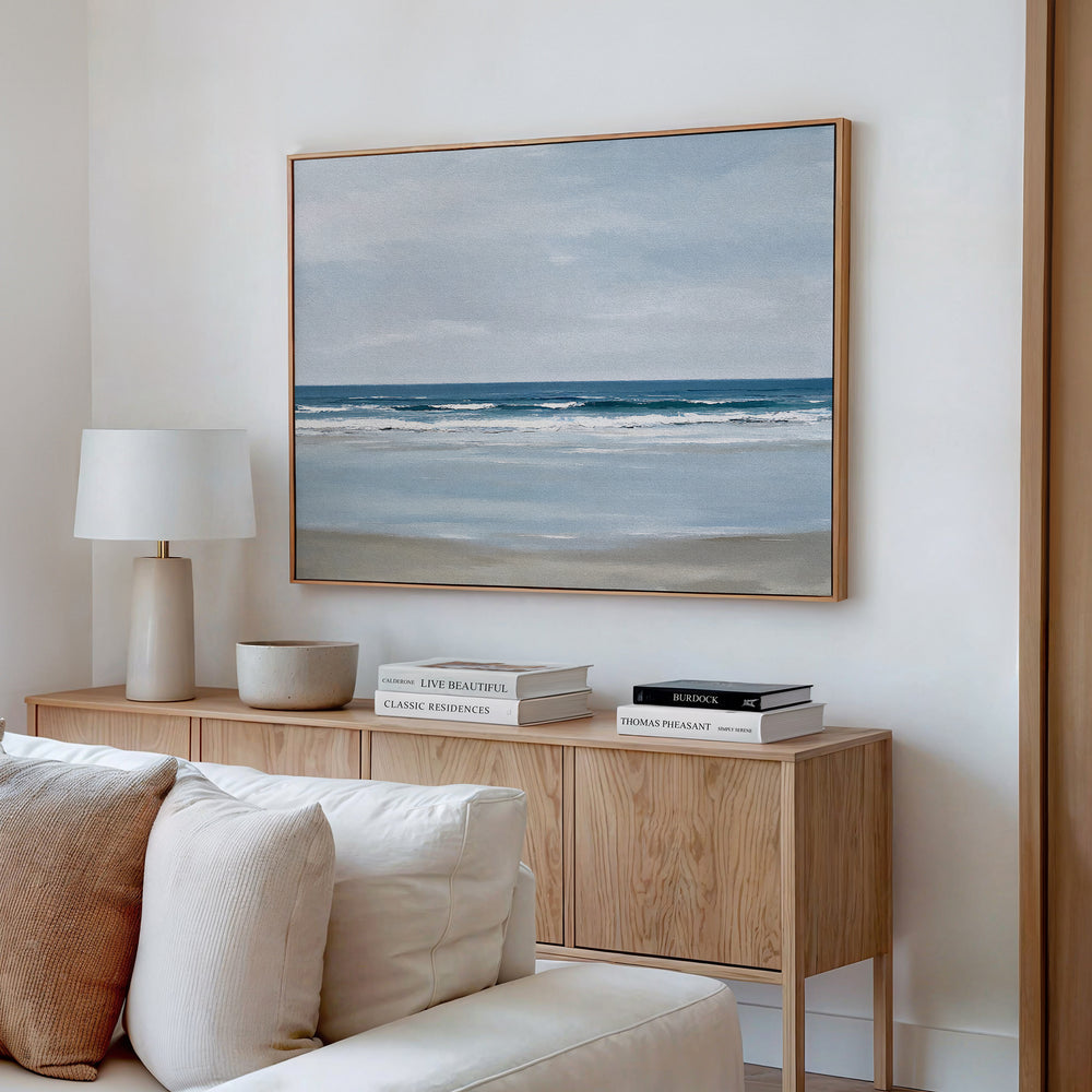 A modern living room features the Soft Tide Canvas Art – Horizontal above a light wood sideboard with books, a lamp, and a bowl. Cream sofa cushions add comfort—making this serene coastal artwork perfect inspiration for bedroom decor.