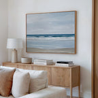 A modern living room features the Soft Tide Canvas Art – Horizontal above a light wood sideboard with books, a lamp, and a bowl. Cream sofa cushions add comfort—making this serene coastal artwork perfect inspiration for bedroom decor.