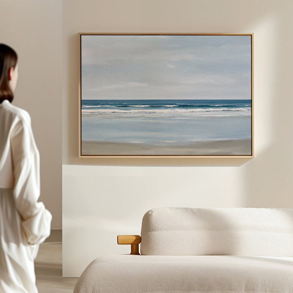 A person in a white shirt stands in a minimal, light room, admiring the Soft Tide Canvas Art – Horizontal—a large framed depiction of calm ocean waves under a cloudy sky—hung above a beige sofa.