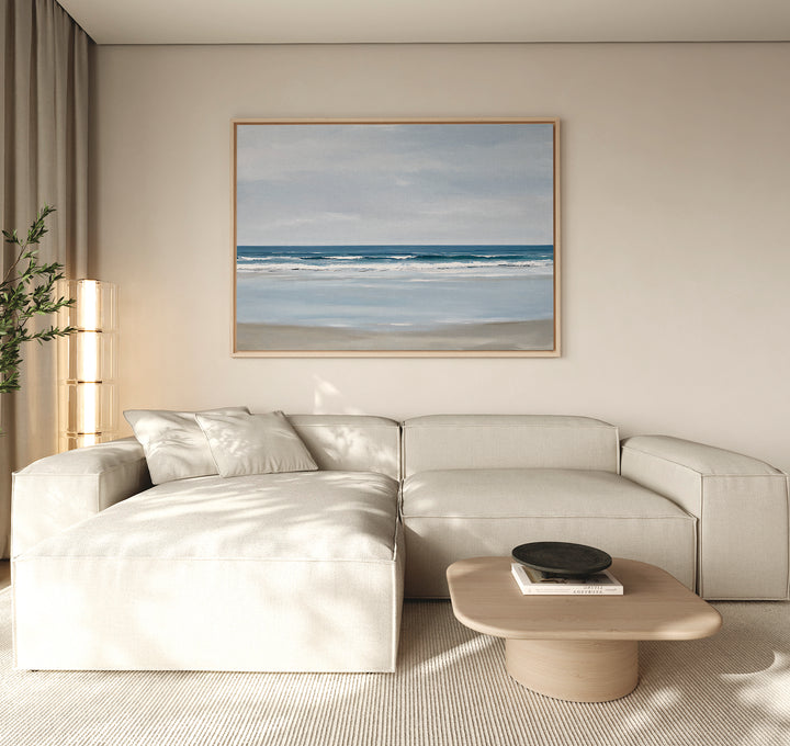 A modern living room with a cream sectional sofa, light wood coffee table, tall plant, and the Soft Tide Canvas Art – Horizontal—a large serene ocean painting—displayed above the sofa.