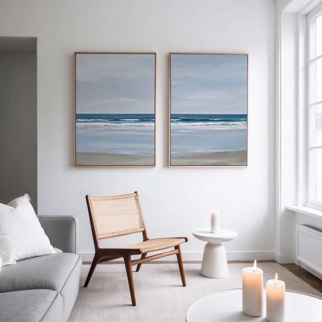 A minimal, bright living room with a light gray sofa, wooden chair, and two lit candles. The white wall displays the Soft Tide Set of 2 coastal beach art prints, while large windows fill the space with natural light.