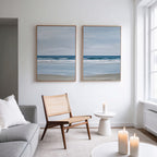 A minimal, bright living room with a light gray sofa, wooden chair, and two lit candles. The white wall displays the Soft Tide Set of 2 coastal beach art prints, while large windows fill the space with natural light.