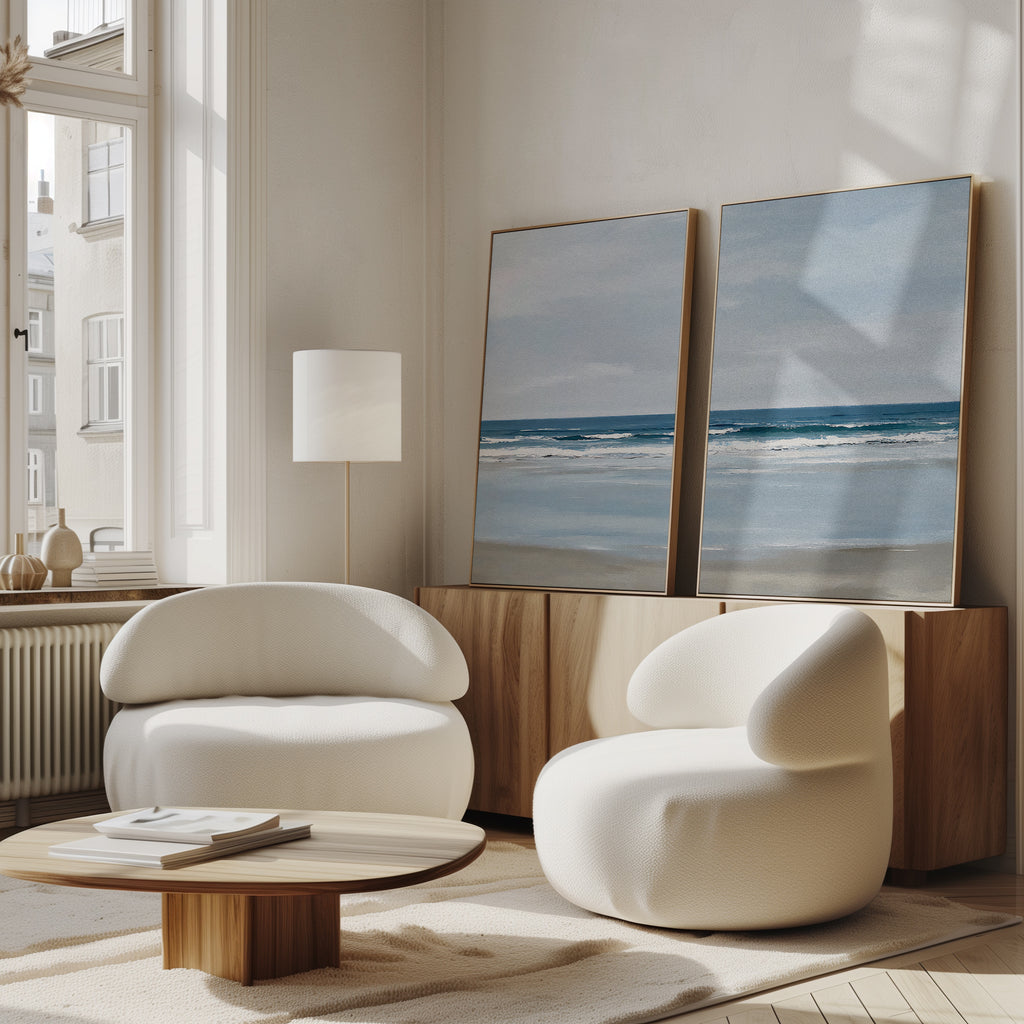 A modern living room features two white rounded armchairs, a wooden coffee table, and the Soft Tide Set of 2—large framed ocean art pieces displayed on a wooden sideboard. Soft natural light pours in from tall windows.