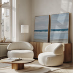 A modern living room features two white rounded armchairs, a wooden coffee table, and the Soft Tide Set of 2—large framed ocean art pieces displayed on a wooden sideboard. Soft natural light pours in from tall windows.