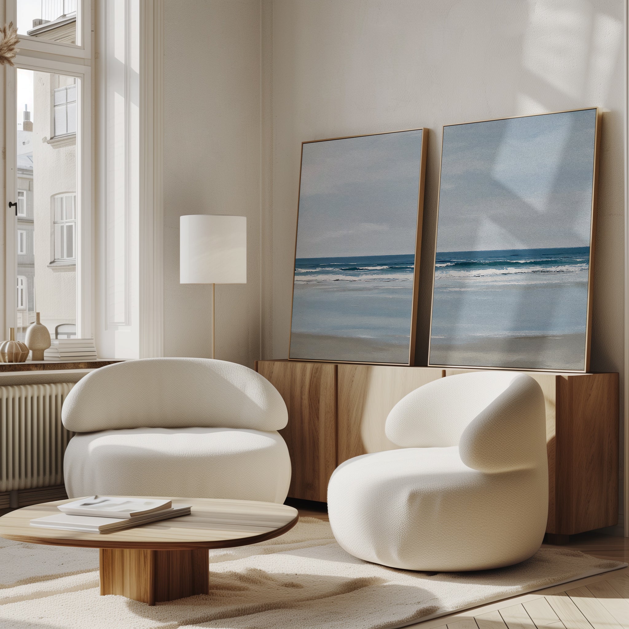 A modern living room features two white rounded armchairs, a wooden coffee table, and the Soft Tide Set of 2—large framed ocean art pieces displayed on a wooden sideboard. Soft natural light pours in from tall windows.