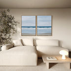 A minimalist living room features a cream sectional sofa, a small wooden coffee table with a lamp and books, a tall plant, and the Soft Tide Set of 2 coastal wall art paintings displayed on the wall.