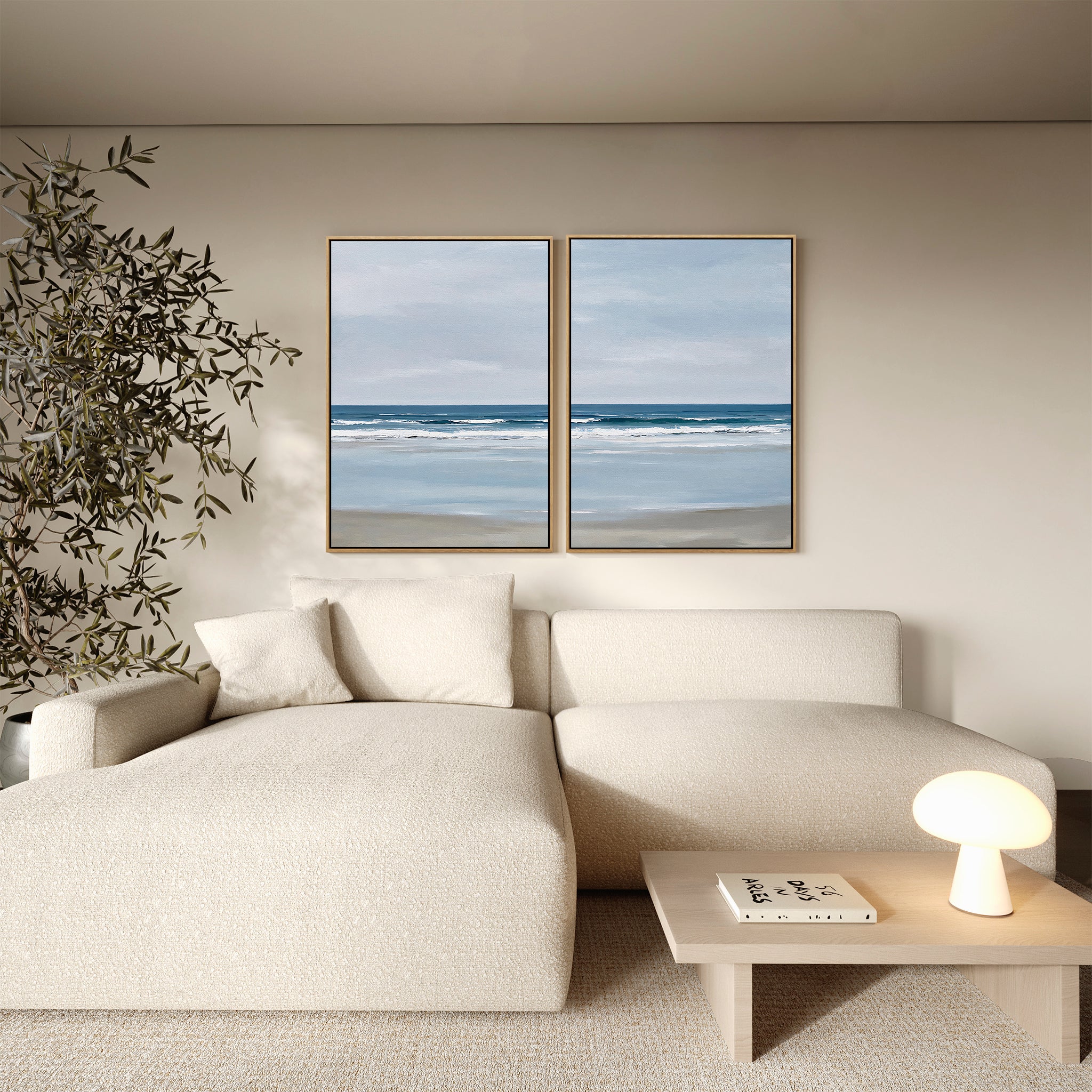 A minimalist living room features a cream sectional sofa, a small wooden coffee table with a lamp and books, a tall plant, and the Soft Tide Set of 2 coastal wall art paintings displayed on the wall.