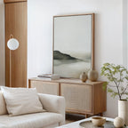Minimalist living room with light wood furniture, a white sofa with cushions, a sideboard holding vases and books, minimalist decor, Soft Valley Canvas Art – Vertical on the wall, a spherical lamp, and a small green plant.