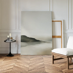 A minimalistic interior showcases the Soft Valley Canvas Art – Vertical resting against a white wall, beside a wooden-frame chair with a white cushion and a small black side table holding candles and books on herringbone wood flooring.