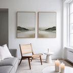 A modern, minimalist living room features the Soft Valley Set of 2 grey sofas, a woven wooden chair, and a white side table with lit candles. Landscape wall art hangs above the chair, creating a peaceful touch as natural light fills the space.