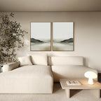 A minimalist living room with a cream sectional, small wooden coffee table with books and a round lamp, a tall plant, and peaceful décor—two framed Soft Valley Set of 2 abstract landscape wall art pieces on a beige wall.