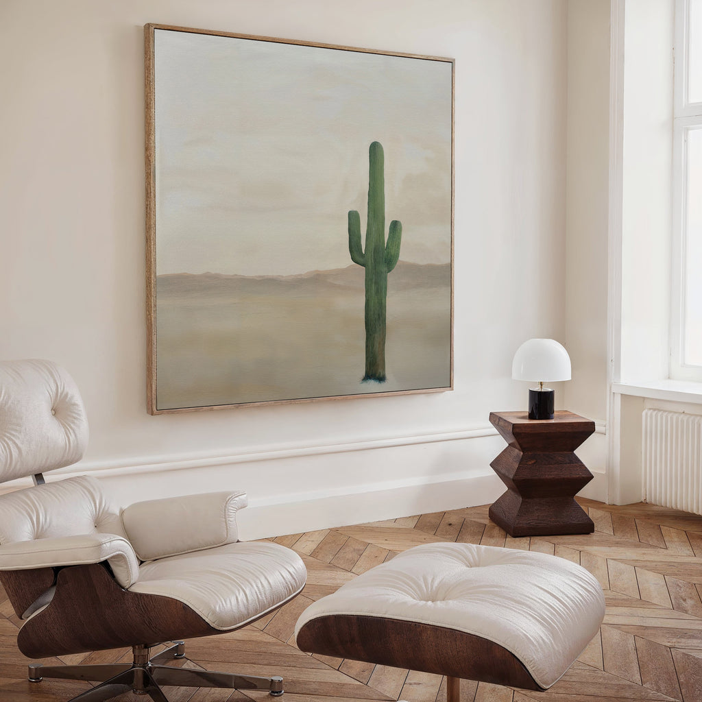 A modern, minimalist living room featuring curated decor: a white lounge chair and ottoman, wooden side table with lamp, and the Solitary Rise Canvas Art—a striking green cactus print on a neutral background.