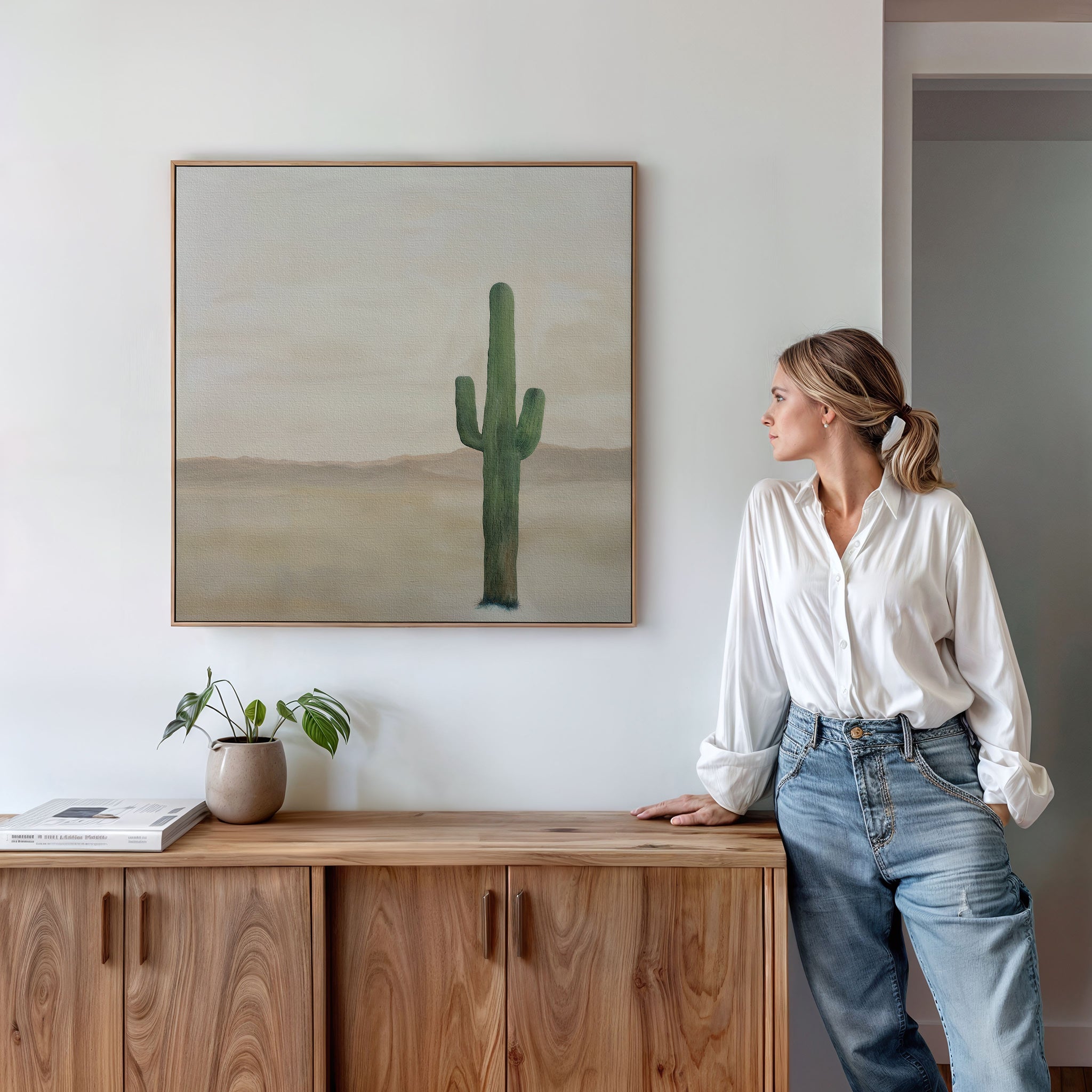 A woman in a white blouse and jeans leans on a wooden cabinet, admiring the Solitary Rise Canvas Art—a minimalist cactus print that adds modern inspiration to the white wall above, ideal for stylish living room decor.