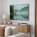 A cozy living space features a wood sideboard with books, lamp, and bowl. Above, the Solitude by the Lake Canvas Art depicts a deer beside reflective water in a tranquil forest. A cream sofa appears partially in the serene foreground.