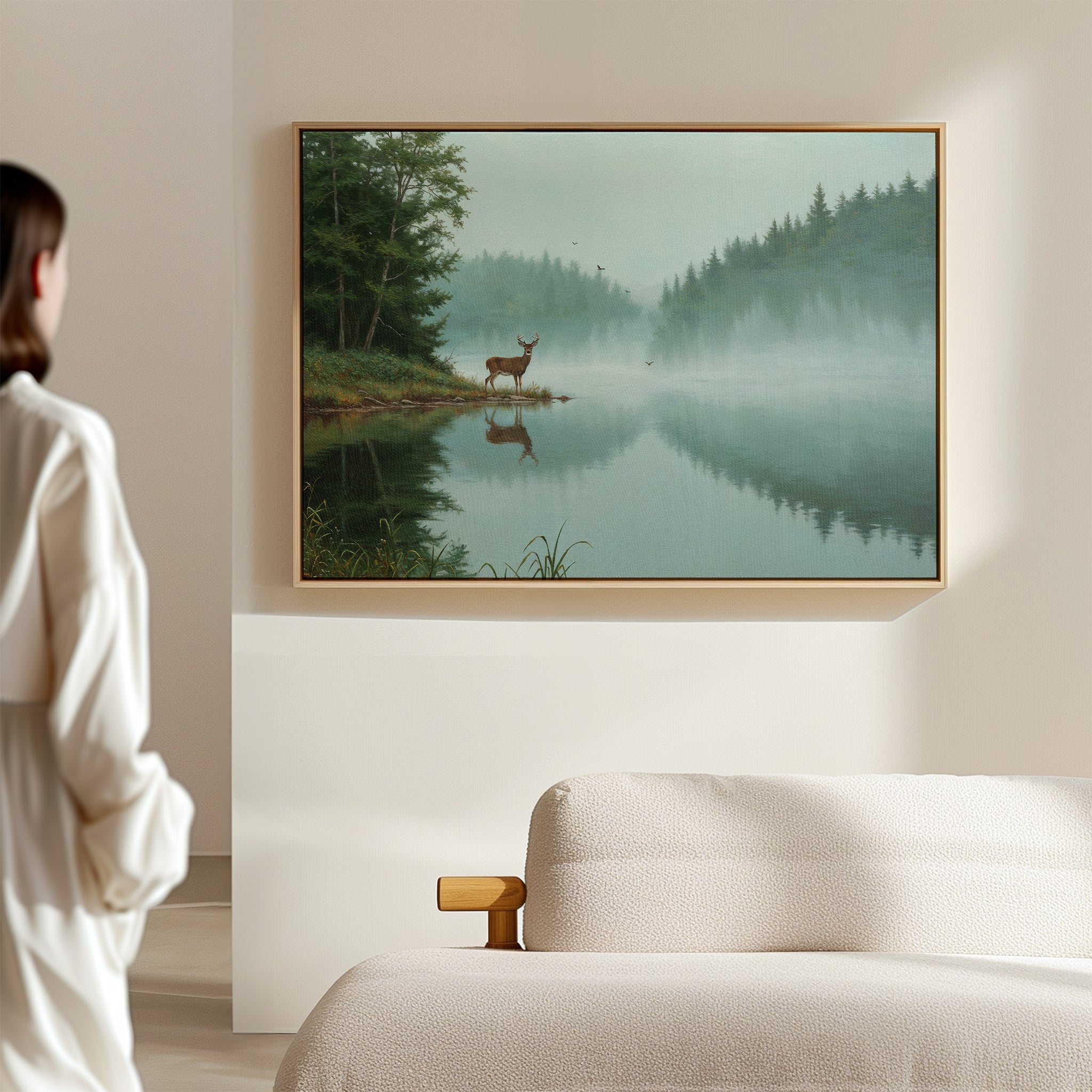 A person stands in a softly lit room, admiring Solitude by the Lake Canvas Art above a beige sofa—a serene scene of a deer by reflective water and evergreens, creating a peaceful forest view.