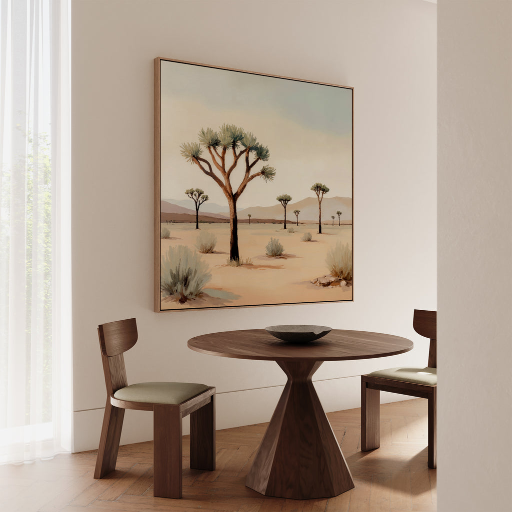 A round wooden table with two chairs sits in a minimalist room. Above, the Sparse Growth Canvas Art depicting Joshua trees hangs on the wall as light seeps through sheer white curtains.