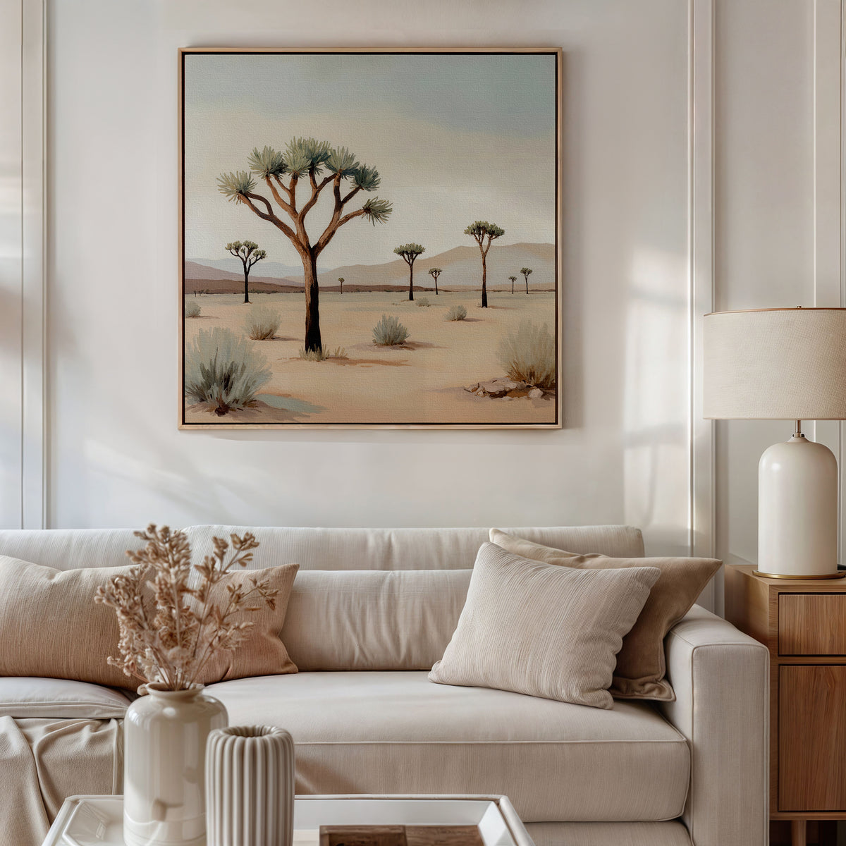 A modern living room with a white sofa, beige cushions, dried flowers in a vase, and minimal decor is enhanced by the Sparse Growth Canvas Art—a framed desert landscape with Joshua trees—creating an inviting, nature-inspired space above the sofa.