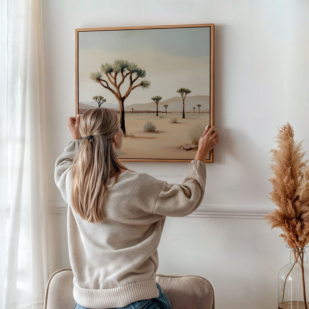 A woman with long blonde hair hangs the Sparse Growth Canvas Art, featuring Joshua trees, on a white wall. Sunlight streams through sheer curtains as dried pampas grass brings a natural touch to the calm, stylish space.