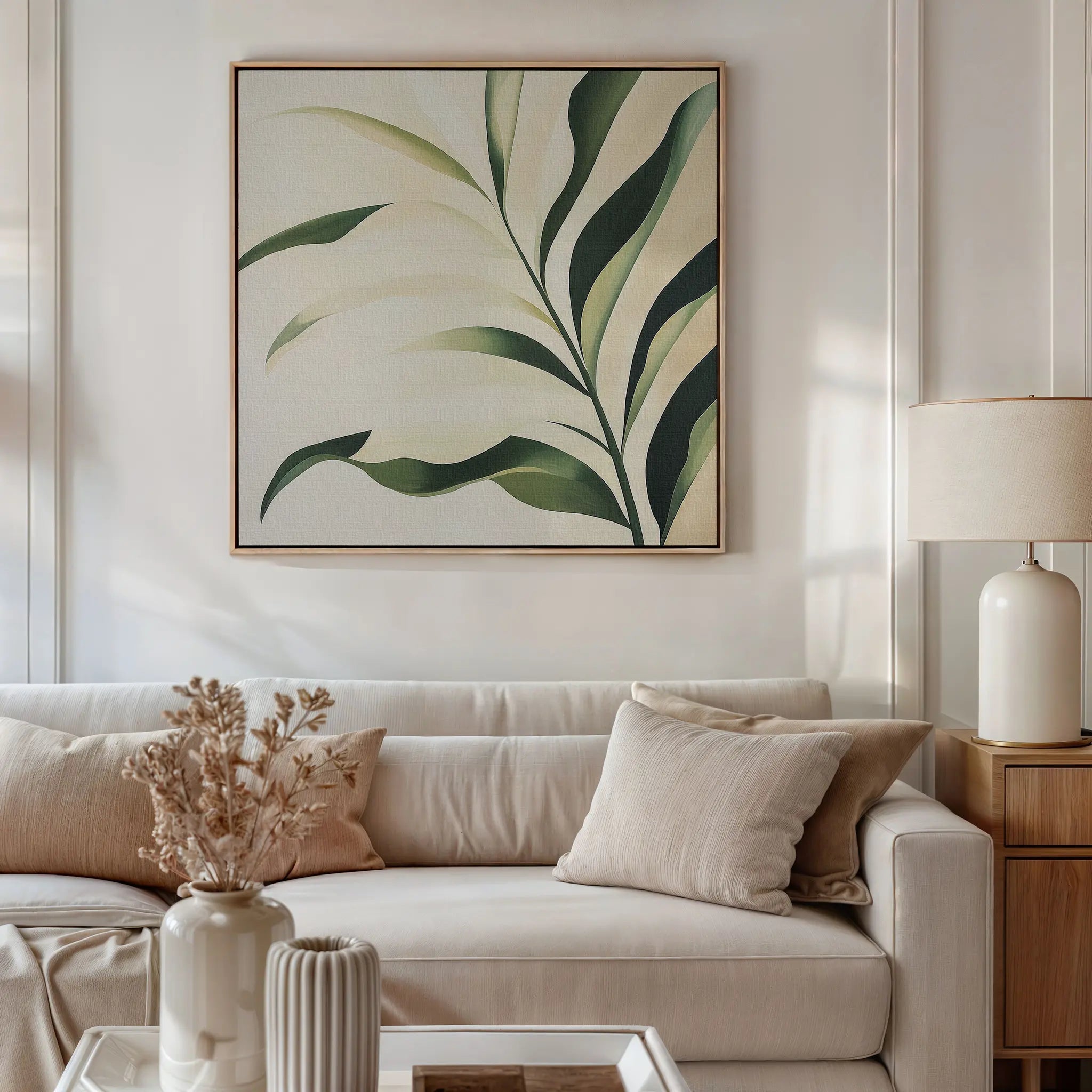 A bright living room with a white sofa, beige and cream cushions, a wooden side table with a ceramic lamp, dried flowers in a vase, and modern decor accented by the Spring Foliage Canvas Art featuring green leaves.