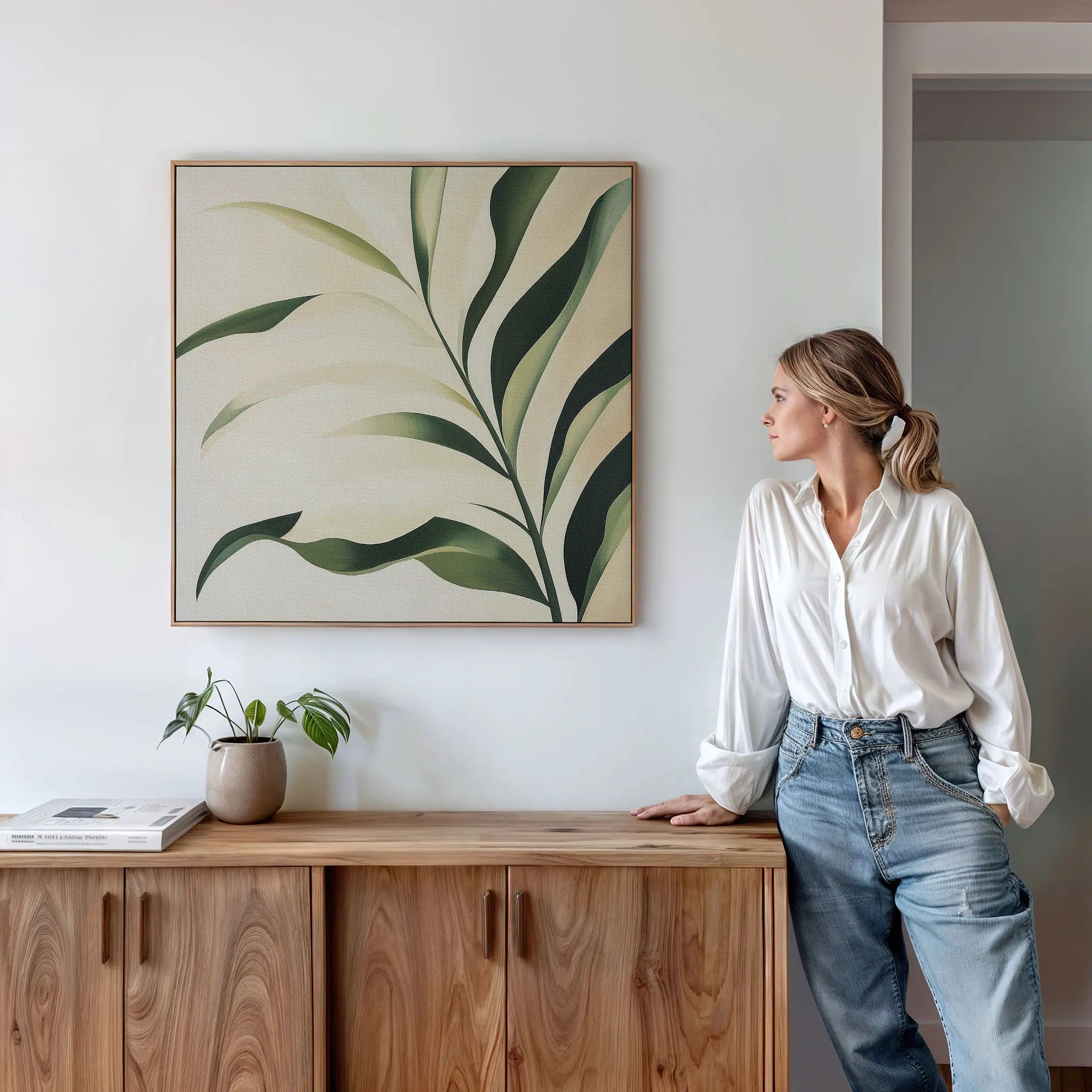 A woman in a white blouse and jeans leans against a wooden cabinet, admiring the Spring Foliage Canvas Art featuring green leaves. A small plant and book on the cabinet add to the stylish, modern interior decor.