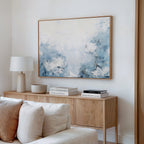 A modern living room with a wooden sideboard, white table lamp, stacked books, and the Spring Hues Canvas Art – Horizontal—a large framed floral painting in soft blue and white tones—hanging above.