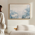 A person in a white shirt stands near a beige sofa, admiring the Spring Hues Canvas Art – Horizontal, which features blue and white floral patterns that add modern style to the living room’s light-colored wall.
