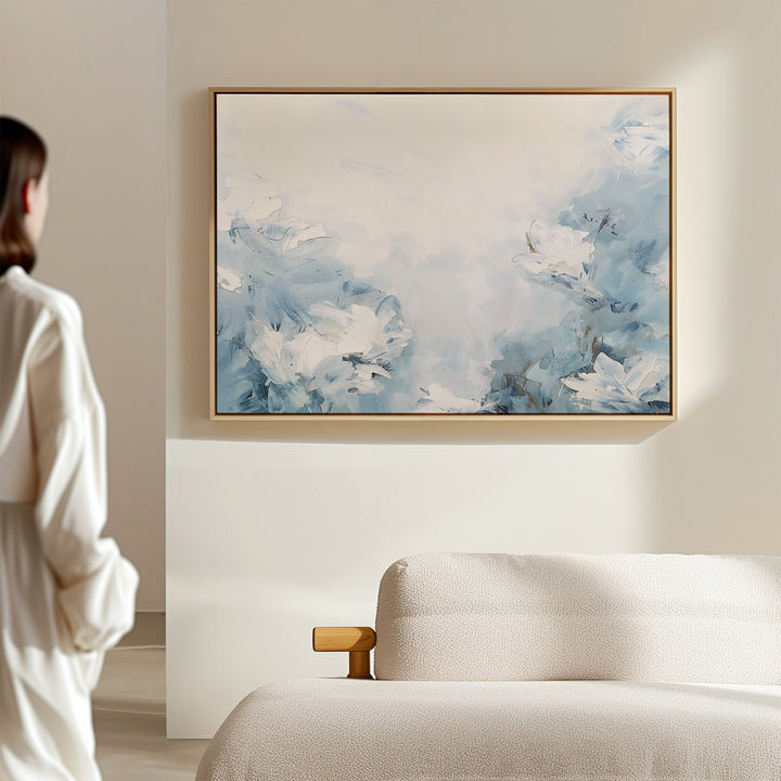 A person in a white shirt stands near a beige sofa, admiring the Spring Hues Canvas Art – Horizontal, which features blue and white floral patterns that add modern style to the living room’s light-colored wall.