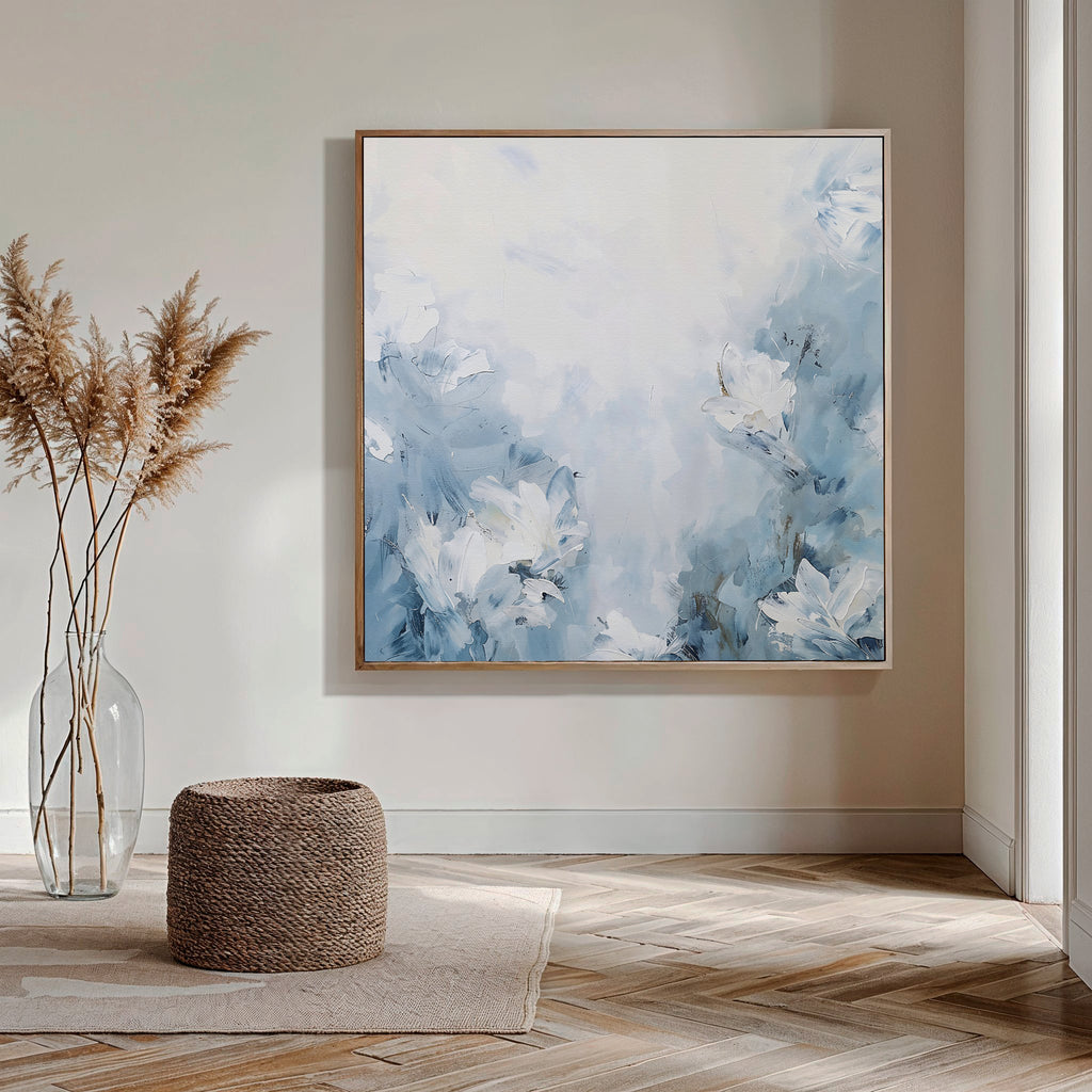 A modern room features Spring Hues Abstract Canvas Art—a large blue and white floral painting—on the wall, complemented by a glass vase with pampas grass, a woven ottoman, wooden herringbone floors, and natural light streaming in from the right.