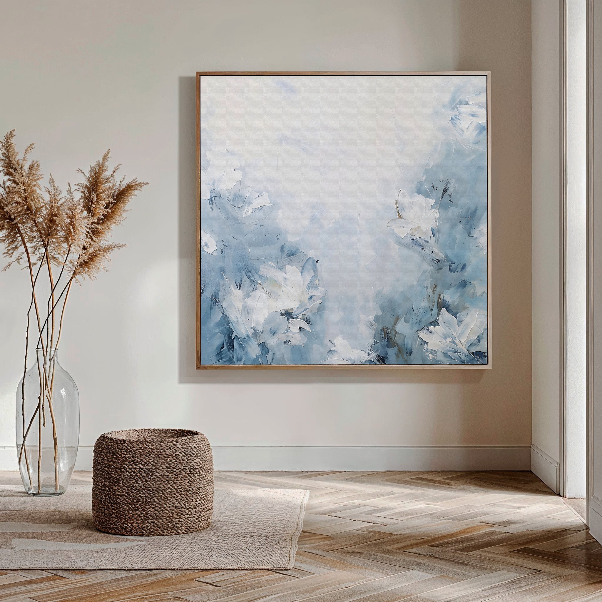 A modern room features Spring Hues Abstract Canvas Art—a large blue and white floral painting—on the wall, complemented by a glass vase with pampas grass, a woven ottoman, wooden herringbone floors, and natural light streaming in from the right.