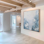 A modern, minimal gallery with light wood beams and white walls features the Spring Hues Abstract Canvas Art—a large blue and white floral painting—prominently displayed as natural light fills the room.