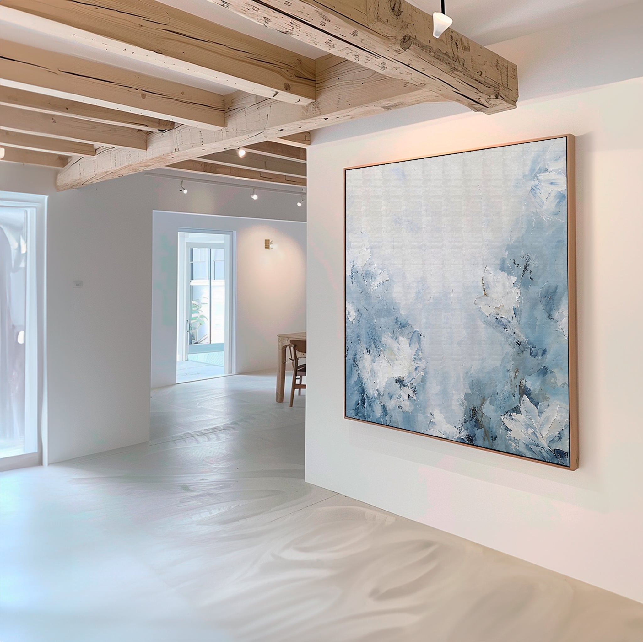 A modern, minimal gallery with light wood beams and white walls features the Spring Hues Abstract Canvas Art—a large blue and white floral painting—prominently displayed as natural light fills the room.