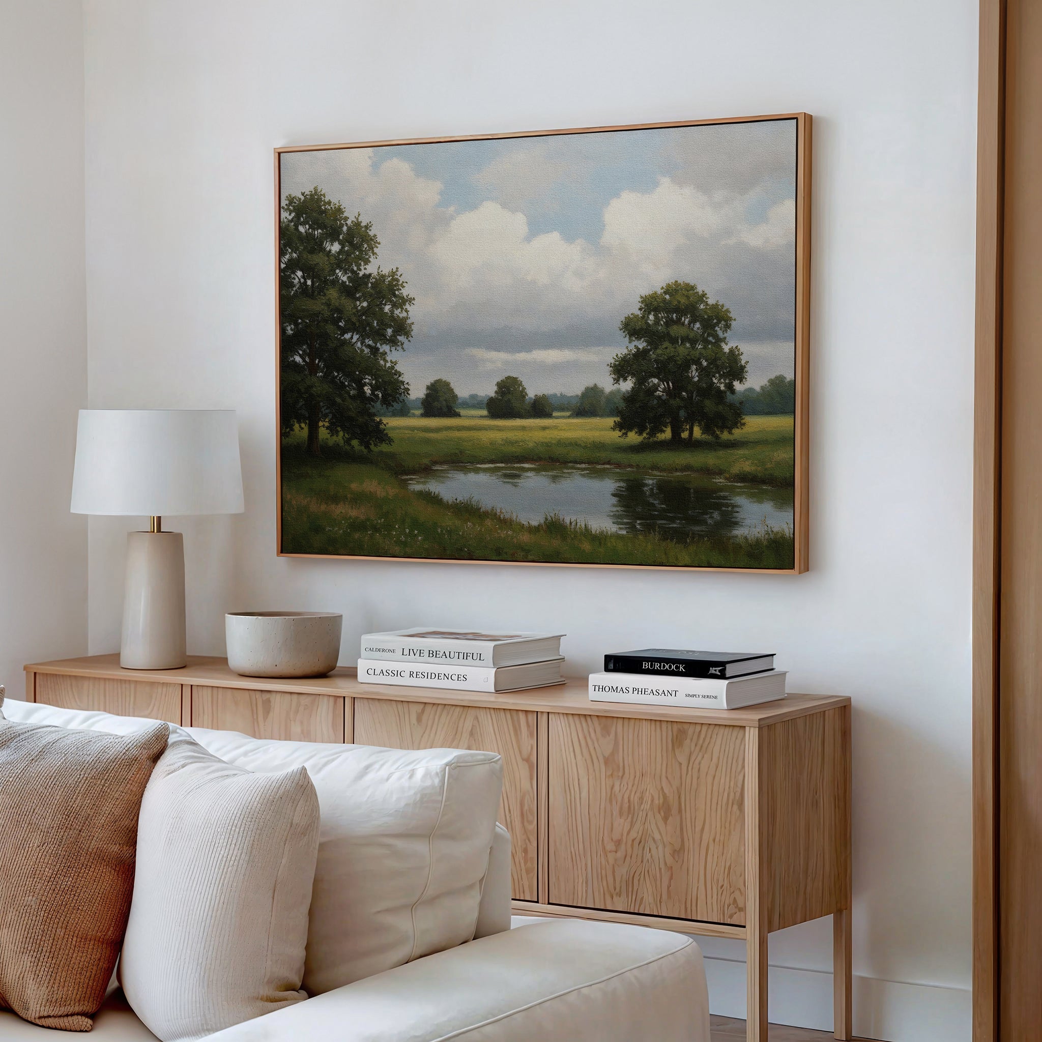 A cozy living room features a light wood sideboard with books and decor, a white table lamp, and the Still Grove Canvas Art of trees and a pond above a cream-colored sofa with cushions.