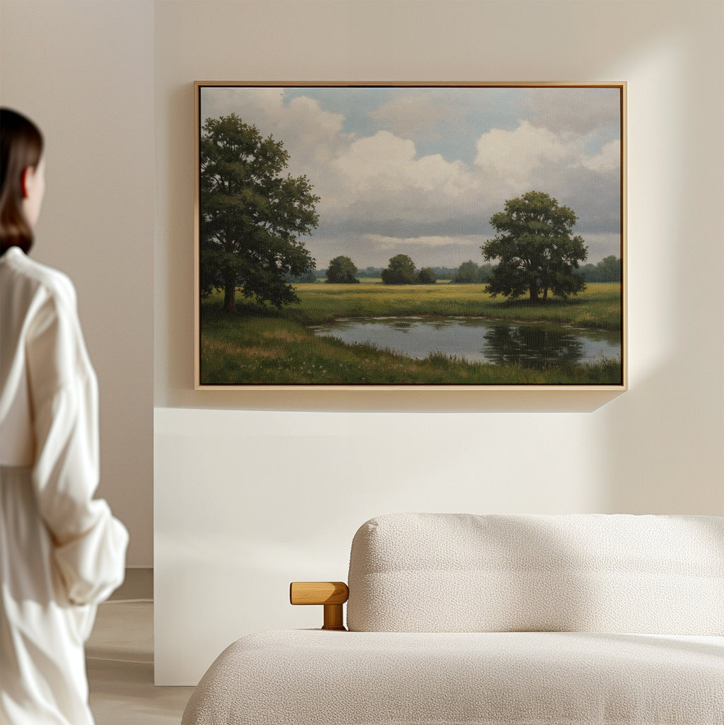 A person stands in a modern, light-colored room admiring the Still Grove Canvas Art, a framed landscape of trees, grass, and a pond displayed above a cream sofa.
