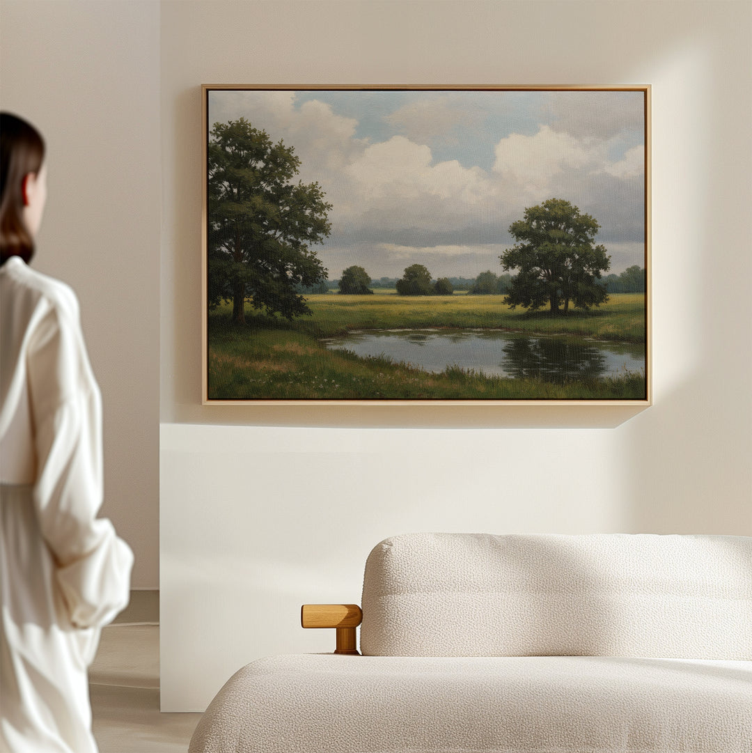 A person stands in a modern, light-colored room admiring the Still Grove Canvas Art, a framed landscape of trees, grass, and a pond displayed above a cream sofa.