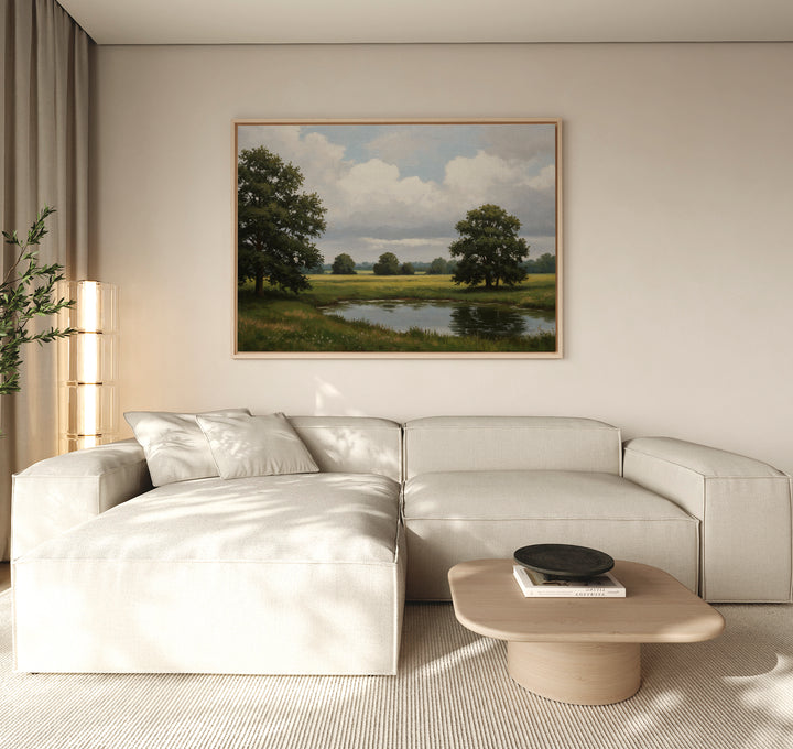 A modern living room with a light beige sectional sofa, wooden coffee table, and the Still Grove Canvas Art—a large landscape painting of trees and a pond—hanging above the sofa. Natural light streams in through curtained windows.