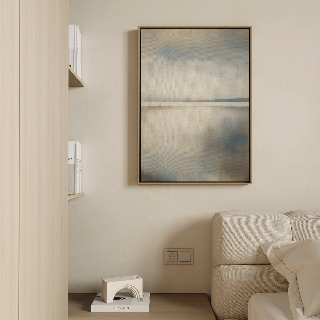 A neutral-toned sofa accents a minimalist living space, complemented by the Still Horizon Canvas Art in soft blue and beige, built-in shelving with books, and a small white decorative object on a wooden surface.