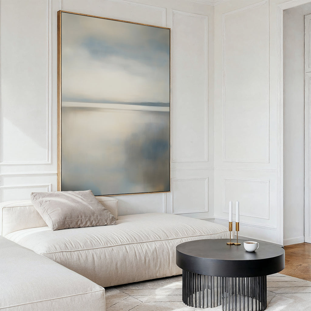 Minimalist living room featuring the Still Horizon Canvas Art, a cream sectional sofa with a beige pillow, round black coffee table, white cup, and two candlesticks—all against white paneled walls and light wood flooring.