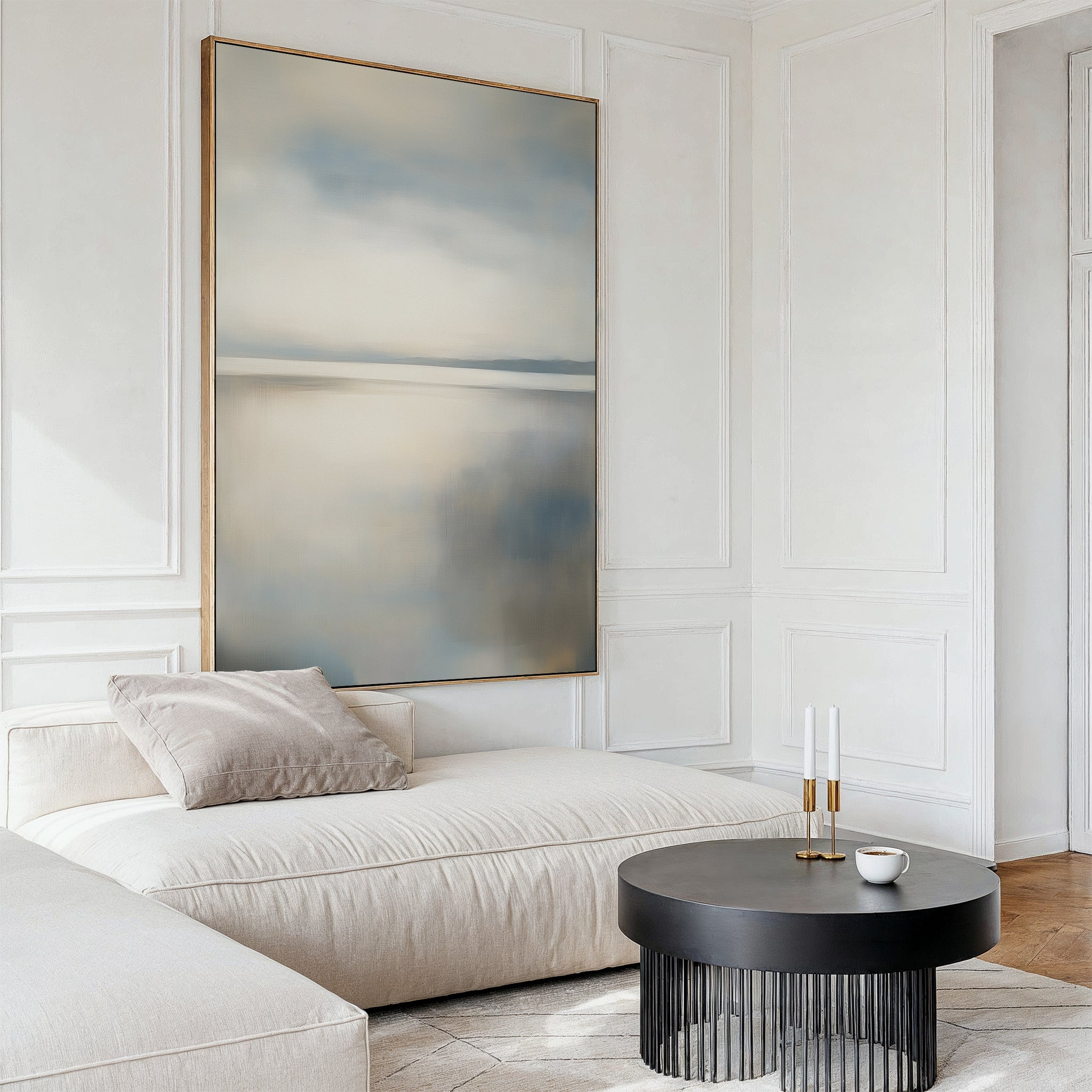 Minimalist living room featuring the Still Horizon Canvas Art, a cream sectional sofa with a beige pillow, round black coffee table, white cup, and two candlesticks—all against white paneled walls and light wood flooring.