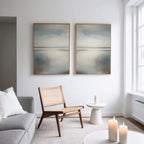 A minimalist living room with a light gray sofa, woven wooden chair, small white side table with a candle, three lit candles on a white coffee table, and the Still Horizon Set of 2 as serene minimalist wall art above.